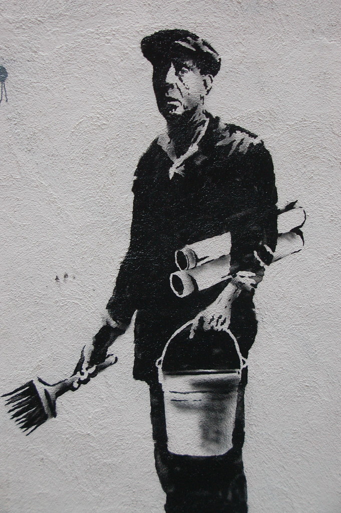 A black and white street mural depicting a worker holding a paintbrush and bucket with rolled-up papers under his arm.
